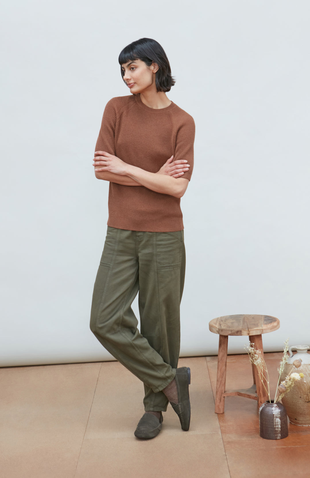women stands in a studio setting wearing a rust-brown short-sleeve knit top by Paul James Knitwear and olive green trousers, paired with dark brown shoes. A small wooden stool and a vase with dried grass are on the floor to the right against a plain off-white wall.
