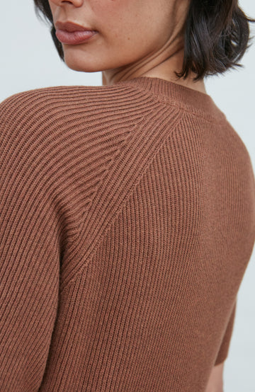 Close-up of a woman wearing a ribbed knit sweater in a warm brown or rust tone, showing the back and shoulder detail against a light neutral background.