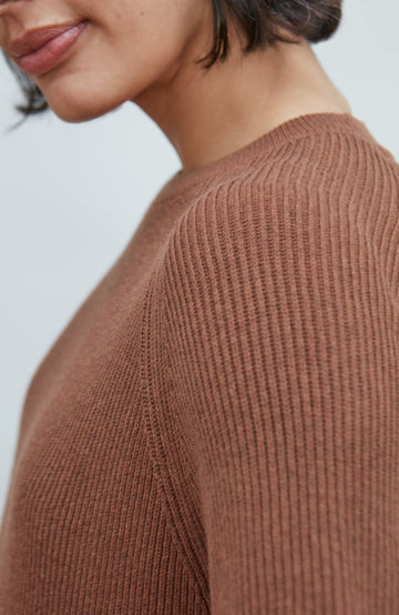 Close-up of a person wearing a ribbed knit sweater in a warm brownish-rose tone against a light gray background.