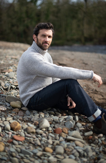 A man sitting on a pebble-strewn beach wearing a light gray turtleneck sweater and dark blue jeans. Made in England with 100% British wool by Paul James Knitwear.