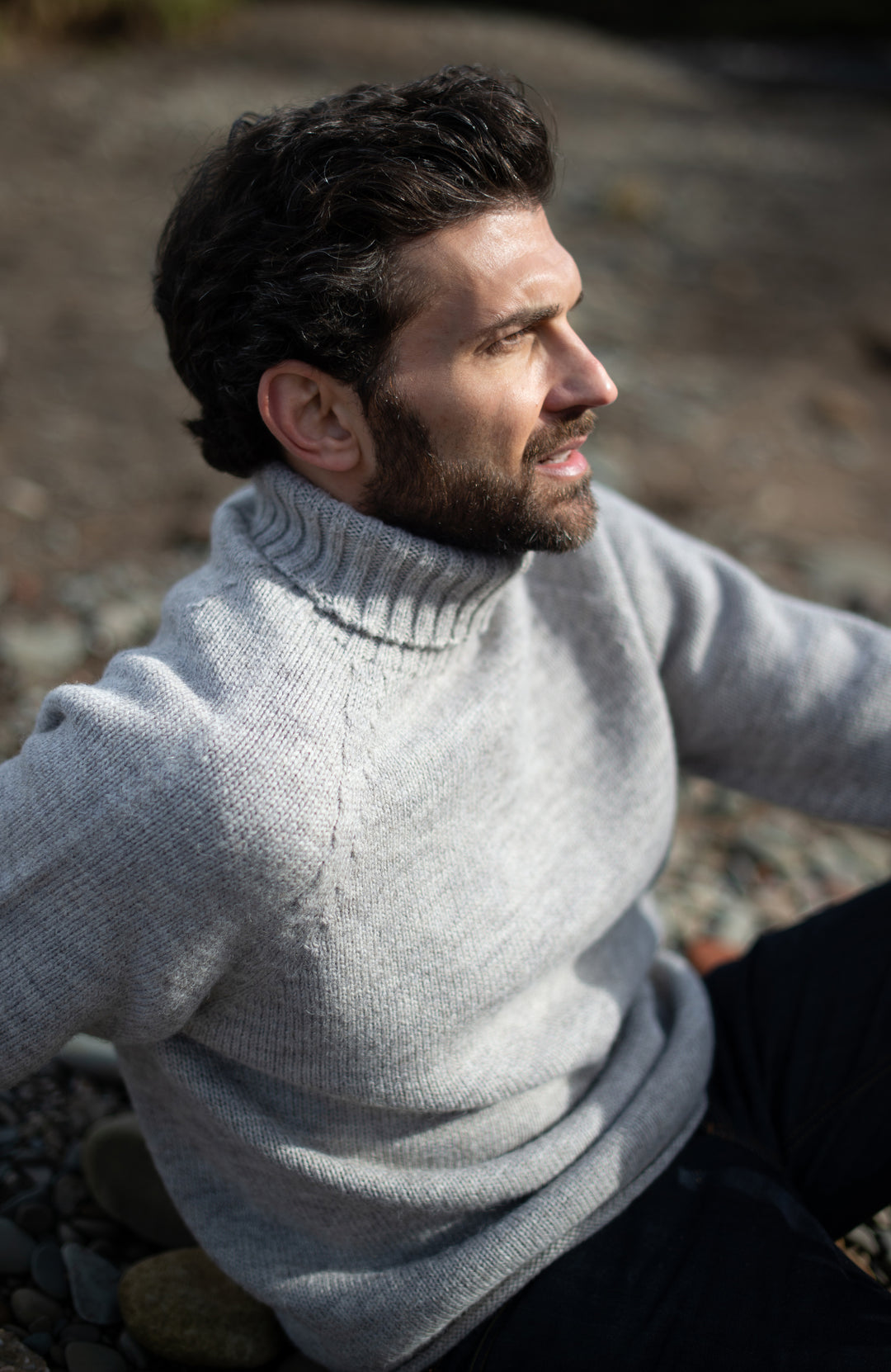 A man wearing a light gray knit turtleneck sweater, sitting outdoors on a rocky surface. Made in England with 100% British wool by Paul James Knitwear. 