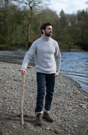 Made in England with 100% British wool by Paul James Knitwear. A man standing on a pebble riverbank wearing a light gray turtleneck sweater, dark jeans, and brown boots, holding a wooden stick with his hand extended.