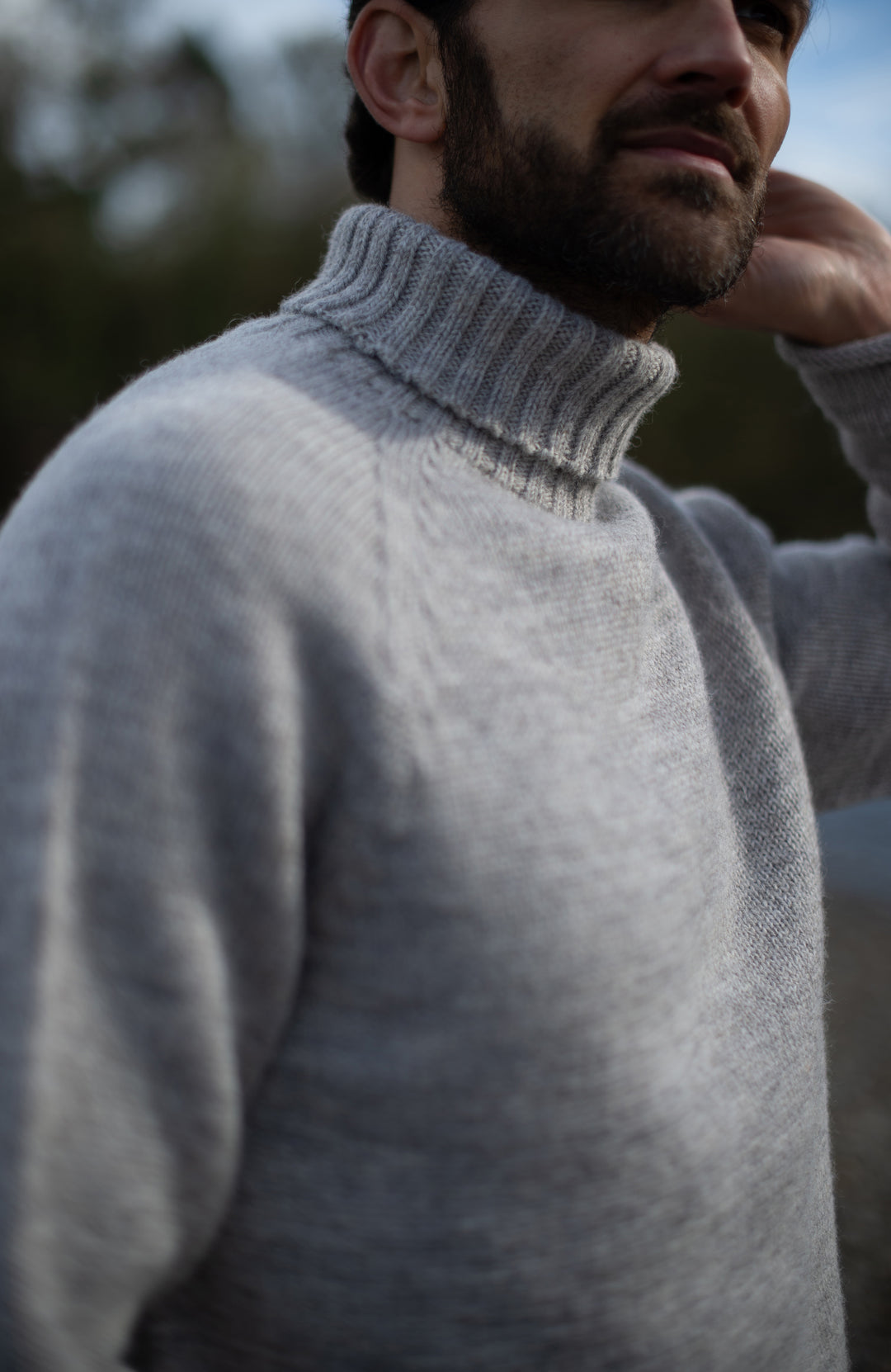 A close-up of a man wearing a light gray Made in England with 100% British wool by Paul James Knitwear. ribbed turtleneck sweater, with a blurred outdoor background.