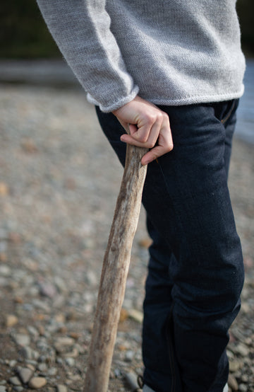 A close-up lifestyle shot of a person wearing a light gray knit sweater Made in England with 100% British wool by Paul James Knitwear and dark blue jeans, holding a rustic wooden walking stick outdoors on a gravel surface.
