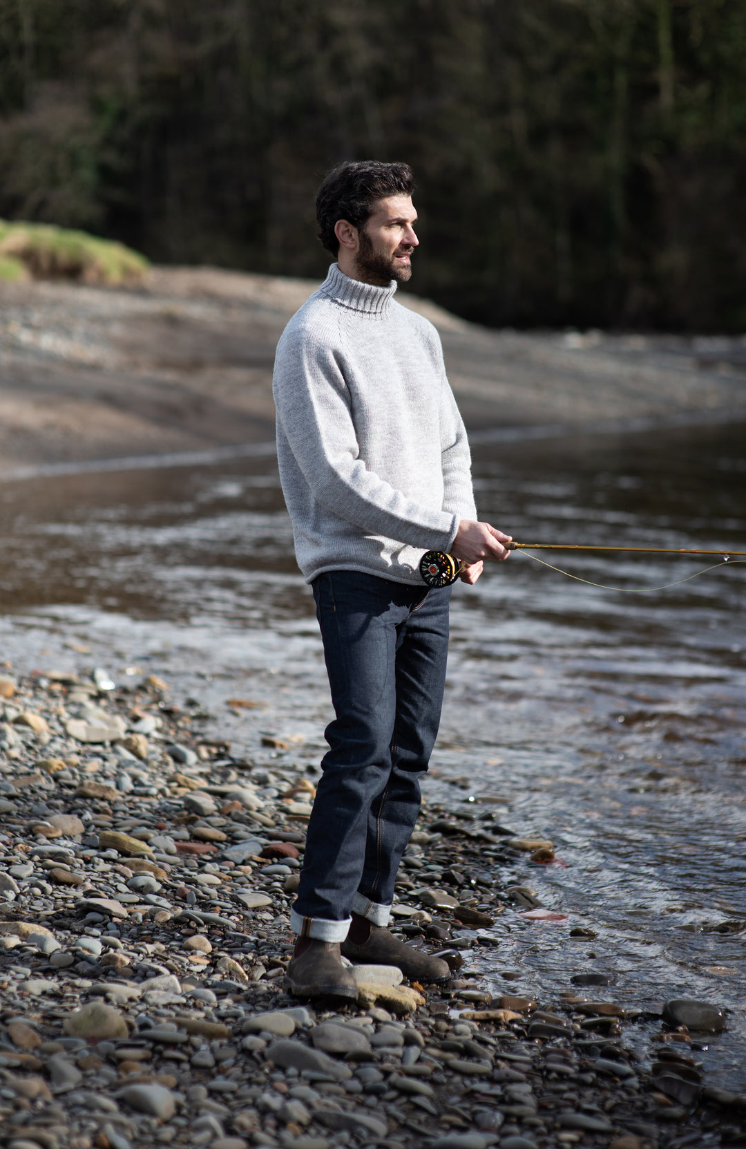 A man stands on a rocky riverbank holding a fly fishing rod, wearing a light gray turtleneck sweater, dark blue jeans, and brown boots. Made in England with 100% British wool by Paul James Knitwear.