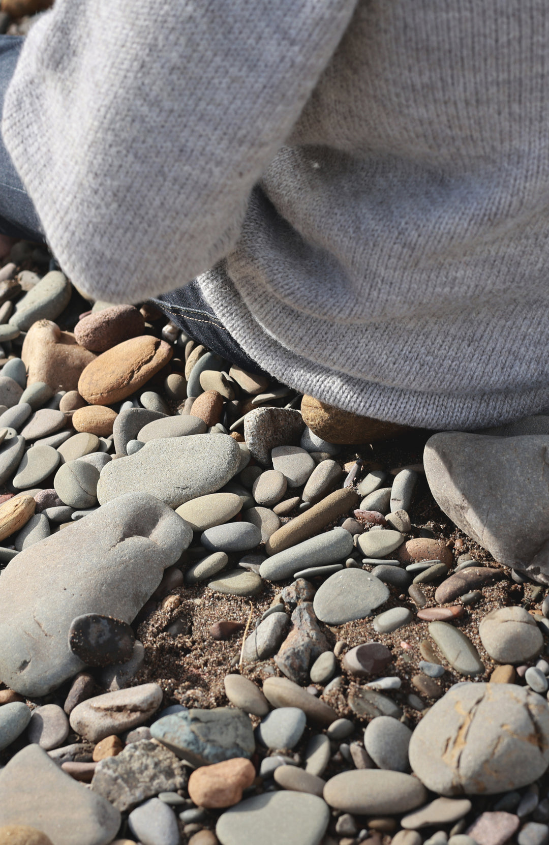 A close-up shot of a person wearing a light gray knit sweater and dark pants, kneeling on a bed of smooth river stones and pebbles. Made in England with 100% British wool by Paul James Knitwear.
