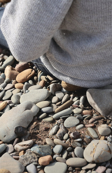 A close-up shot of a person wearing a light gray knit sweater and dark pants, kneeling on a bed of smooth river stones and pebbles. Made in England with 100% British wool by Paul James Knitwear.