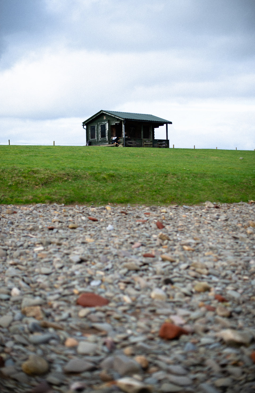 A small dark cabin with a porch sits on a grassy hill under a cloudy sky, with a gravel foreground in the foreground.