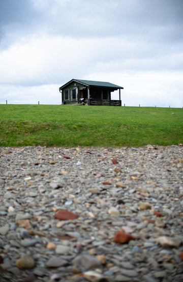 A small dark cabin with a porch sits on a grassy hill under a cloudy sky, with a gravel foreground in the foreground.