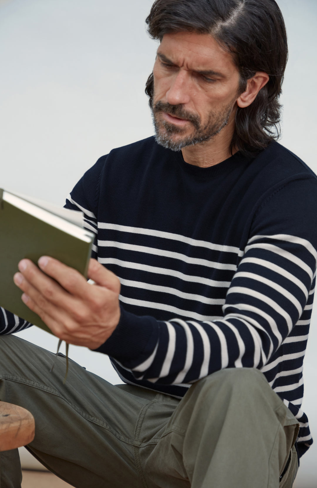 A man with long dark hair and a beard sits reading a book, wearing a navy and white horizontally striped long-sleeve cotton sweater by Paul James Knitwear and olive green pants against a light gray background.