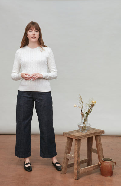 Woman wearing a white cable sweater and dark jeans standing next to a wooden stool with a vase on it.