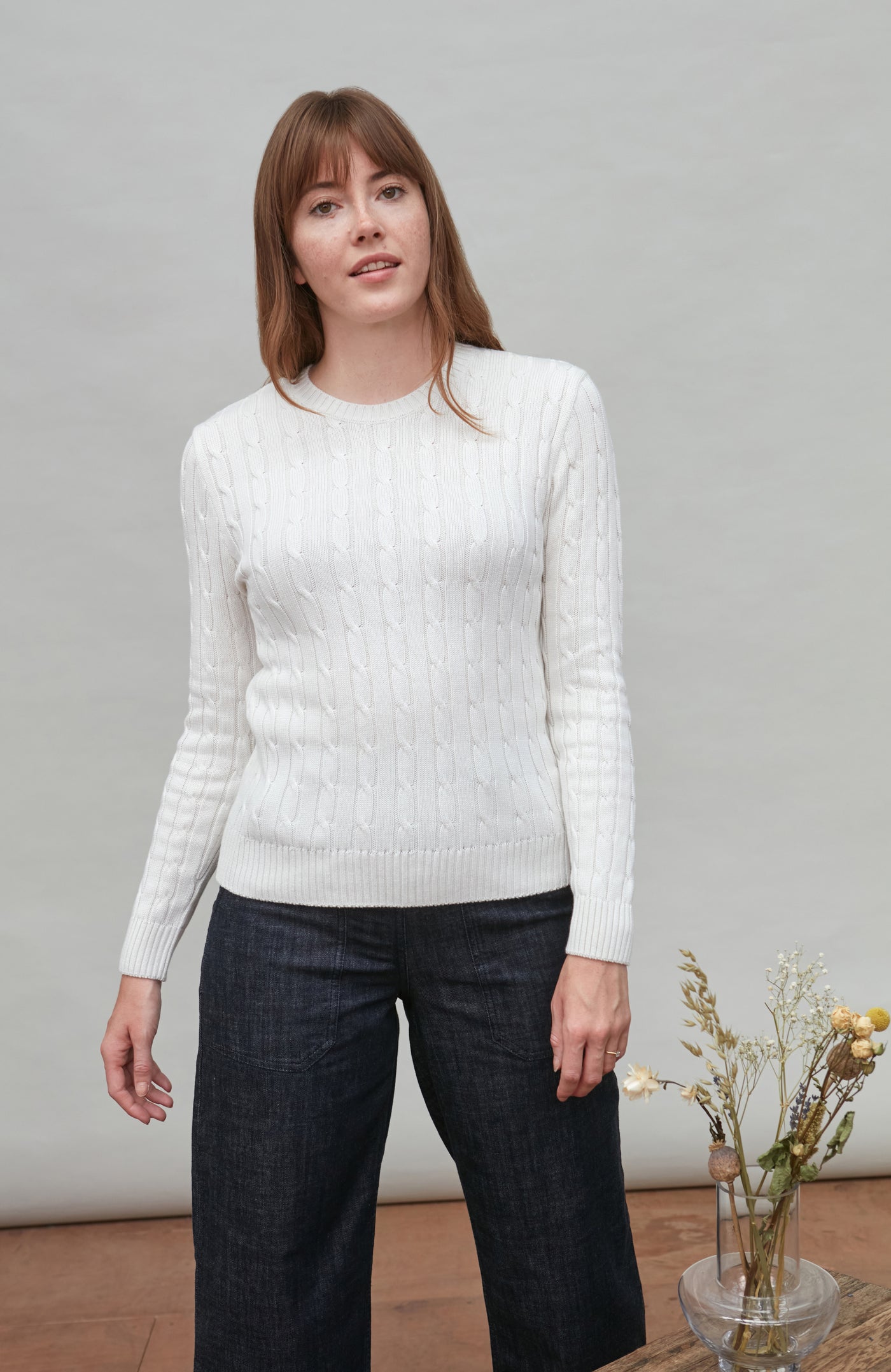 Woman wearing a white cable sweater and dark plaid pants against a plain background