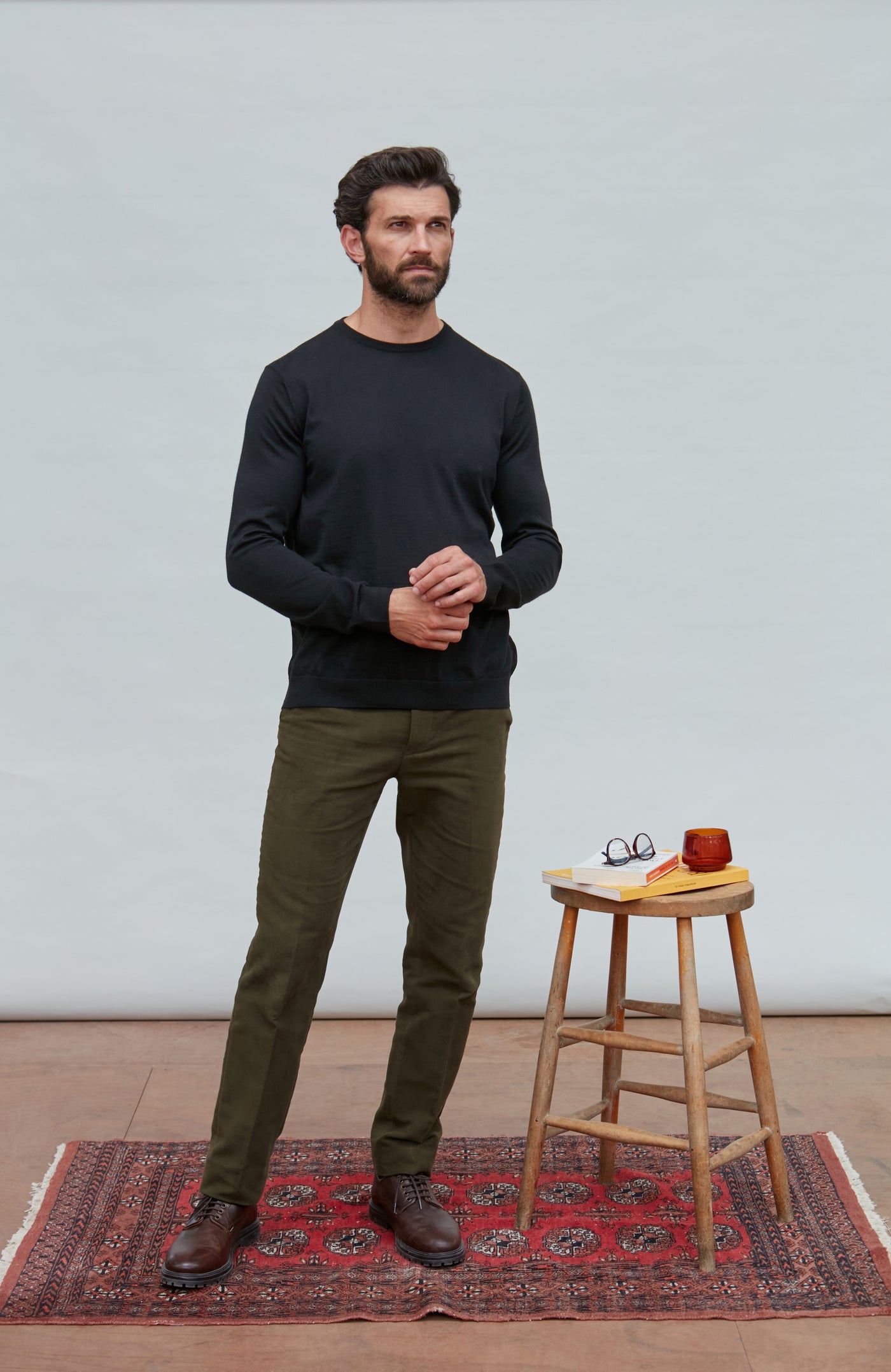 Man wearing a black merino silk sweater and green pants standing on a red patterned rug with a stool and small items in the background.