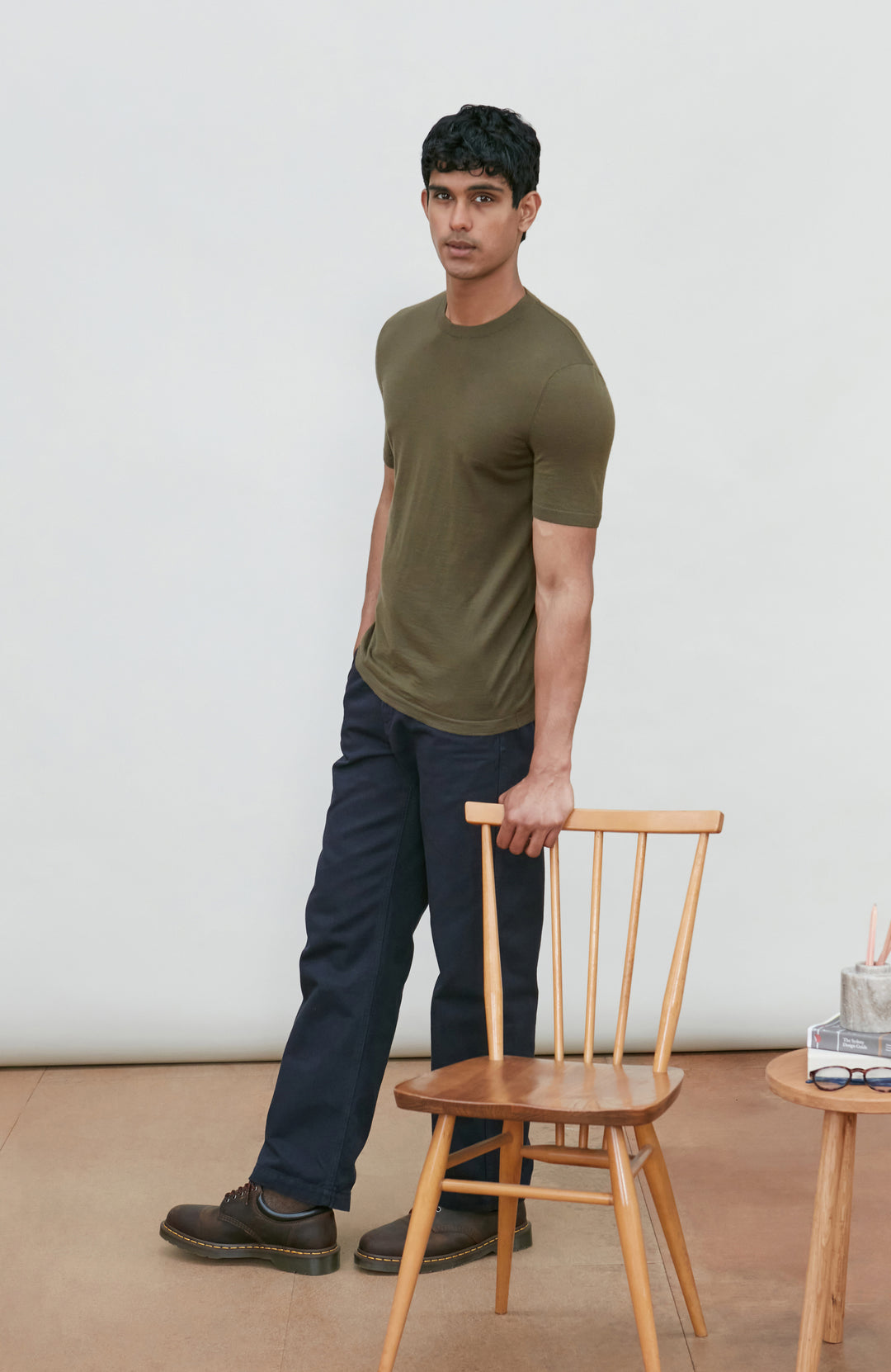 A man wearing a fitted short-sleeve crew-neck merino wool t-shirt in olive green by Paul James Knitwear and dark navy pants stands in a studio setting, holding the back of a light wood chair.