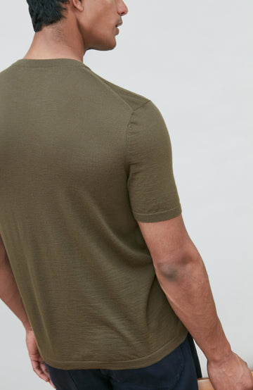 A man wearing a fitted short-sleeve crew-neck merino wool t-shirt in olive green by Paul James Knitwear, shown from a side-back angle against a plain light gray background.