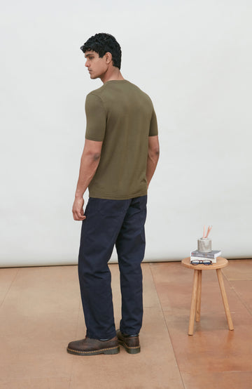 A man wearing a fitted short-sleeve crew-neck merino wool t-shirt in olive green by Paul James Knitwear and dark navy pants, standing in a minimalist studio with a light-colored wall and terracotta floor. A small wooden stool with a cup is on the right.