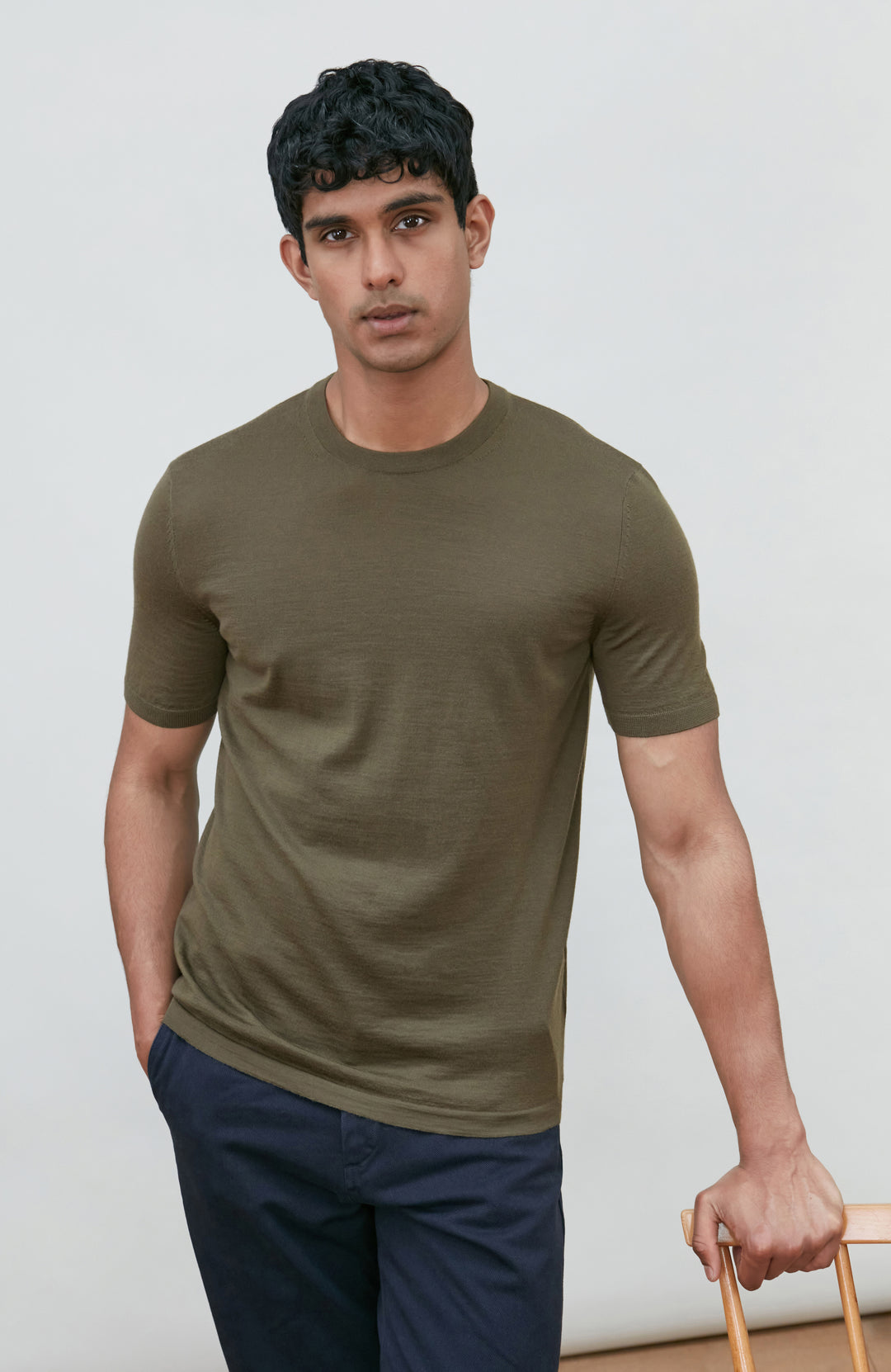 A man wearing a fitted short-sleeve crew-neck merino wool t-shirt in olive green by Paul James Knitwear, paired with navy blue pants, standing against a light gray studio background.