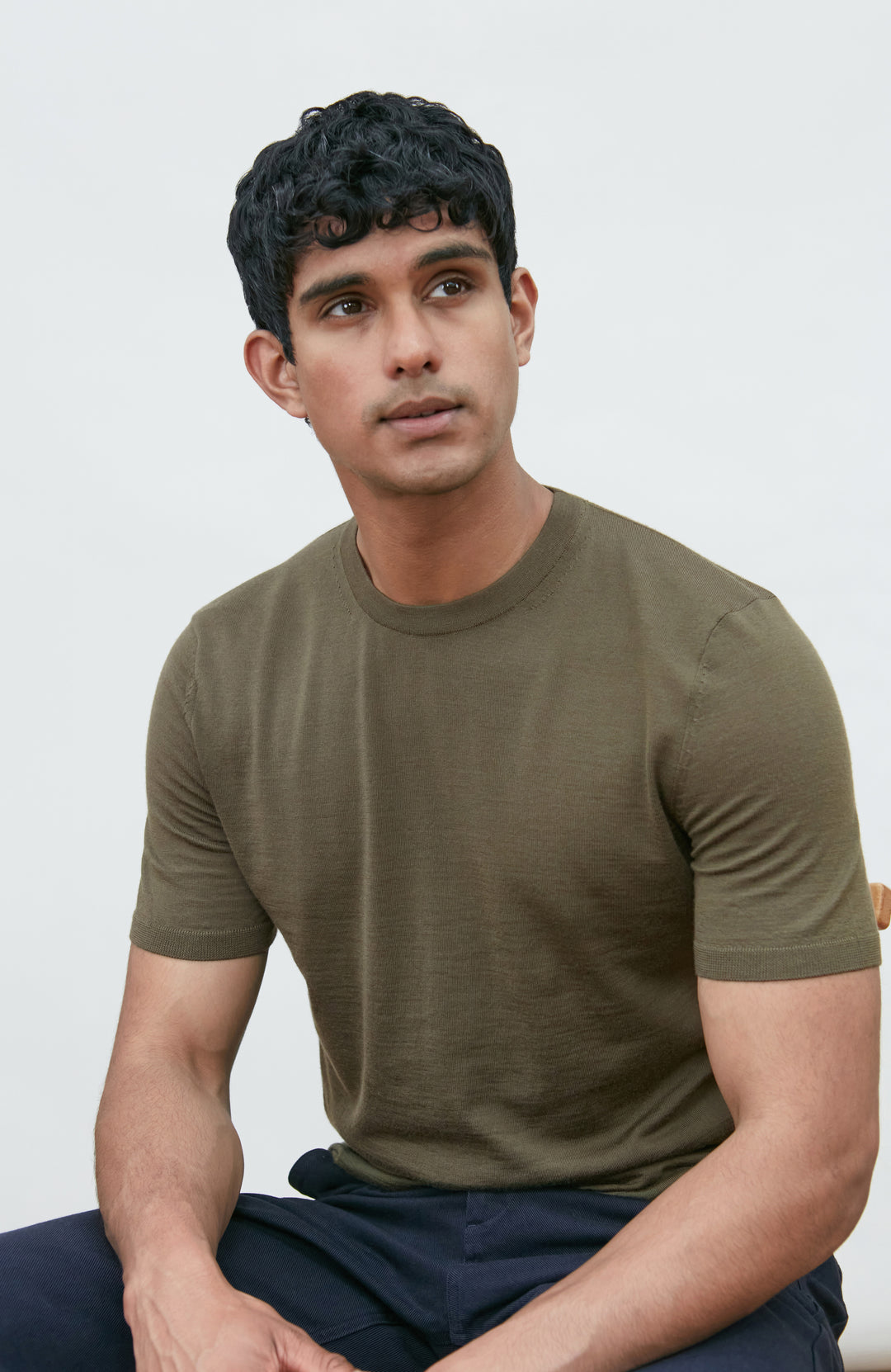 A man wearing a fitted short-sleeve crew-neck merino wool t-shirt in olive green by Paul James Knitwear, paired with dark navy pants, seated against a plain light gray background.
