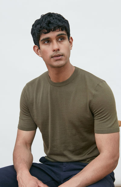 A man wearing a fitted short-sleeve crew-neck merino wool t-shirt in olive green by Paul James Knitwear, paired with dark navy pants, seated against a plain light gray background.