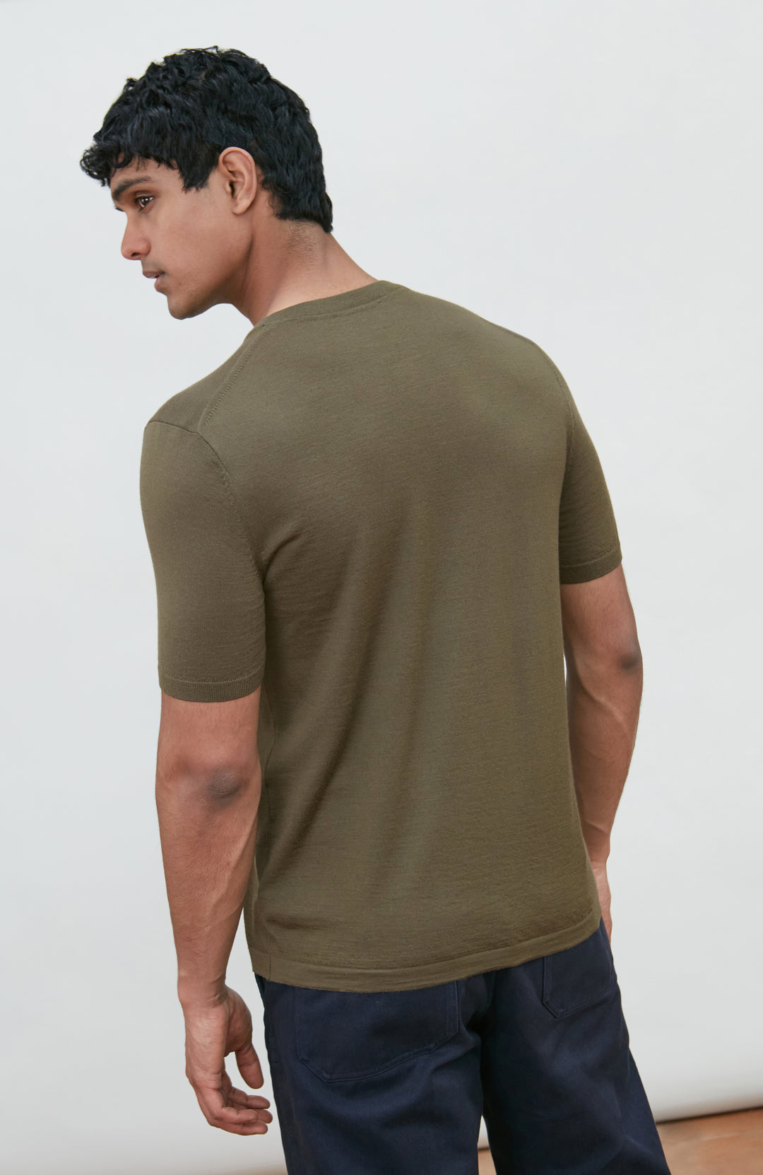 A man wearing a fitted short-sleeve crew-neck merino wool t-shirt in olive green by Paul James Knitwear, shown from a three-quarters back view against a plain off-white background.