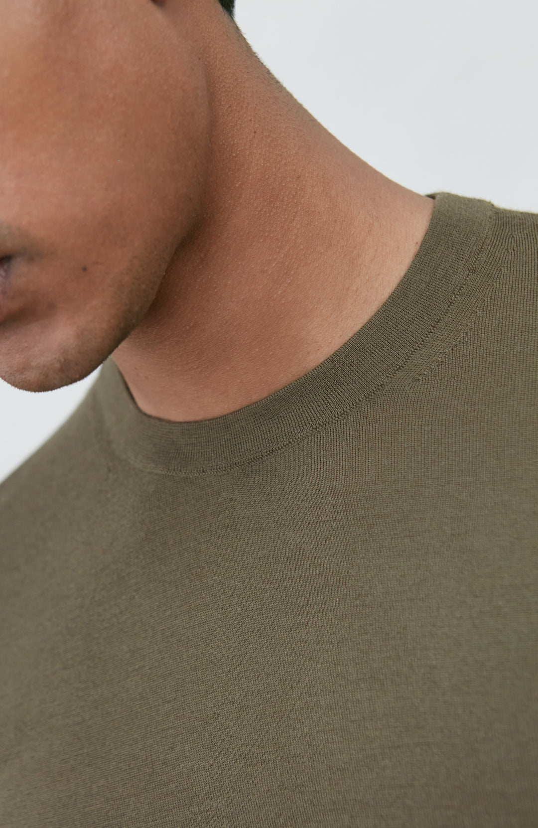 Close-up of a person wearing a ribbed crew-neck t-shirt in olive green, showing the collar and upper chest against a light gray background.