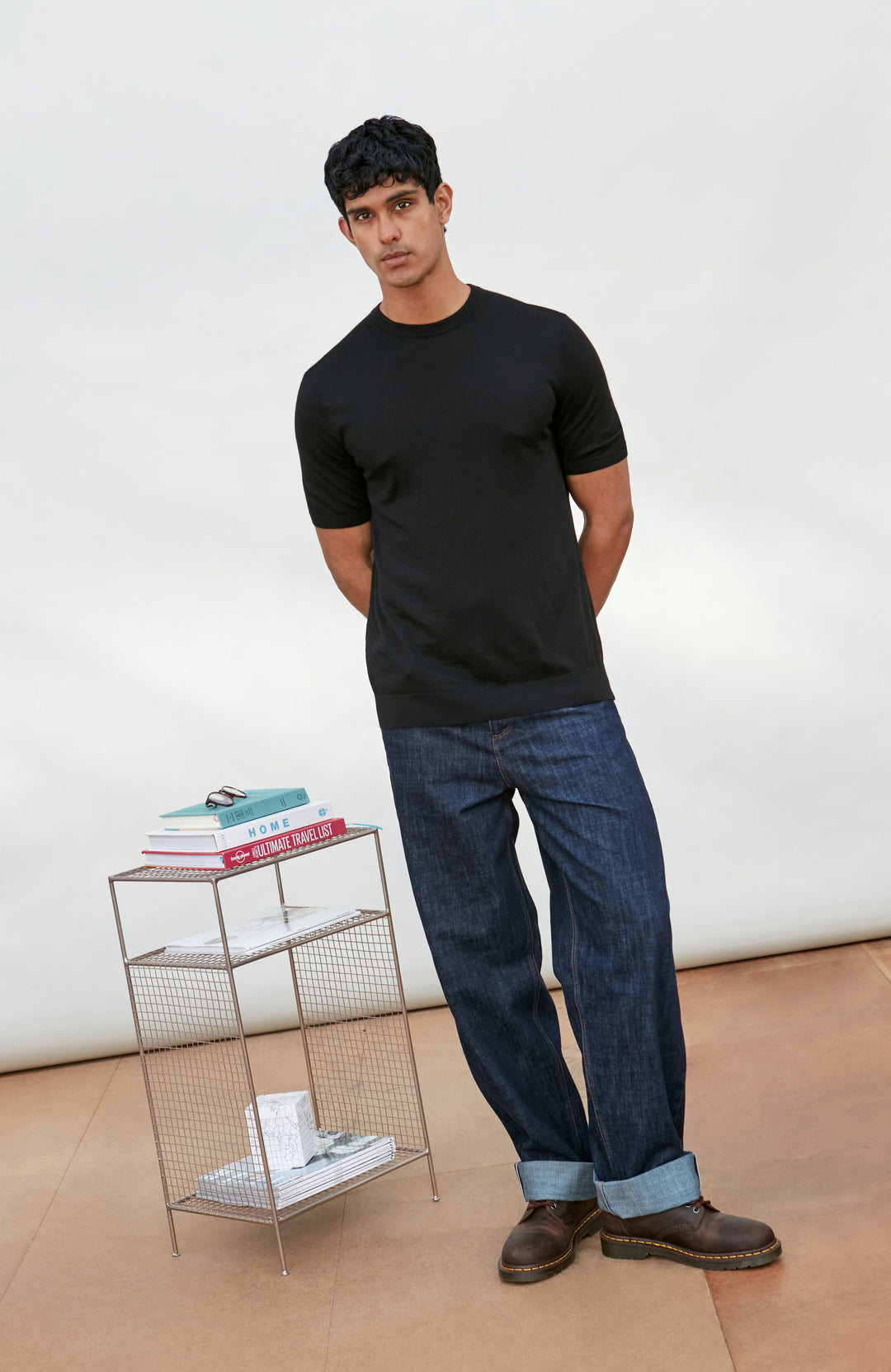 A man wearing a black short-sleeve crewneck knit sweater by Paul James Knitwear and blue jeans, standing against a light off-white background.
