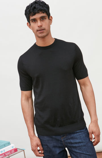 A man wearing a black short-sleeve crewneck knit sweater by Paul James Knitwear and blue jeans, standing against a light off-white background.