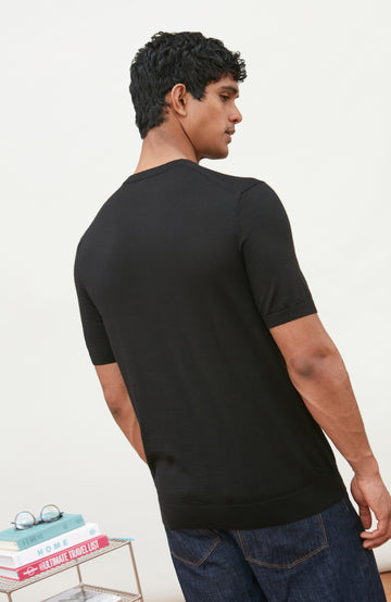 A man wearing a black short-sleeve crewneck knit sweater by Paul James Knitwear and blue jeans, standing against a light off-white background.