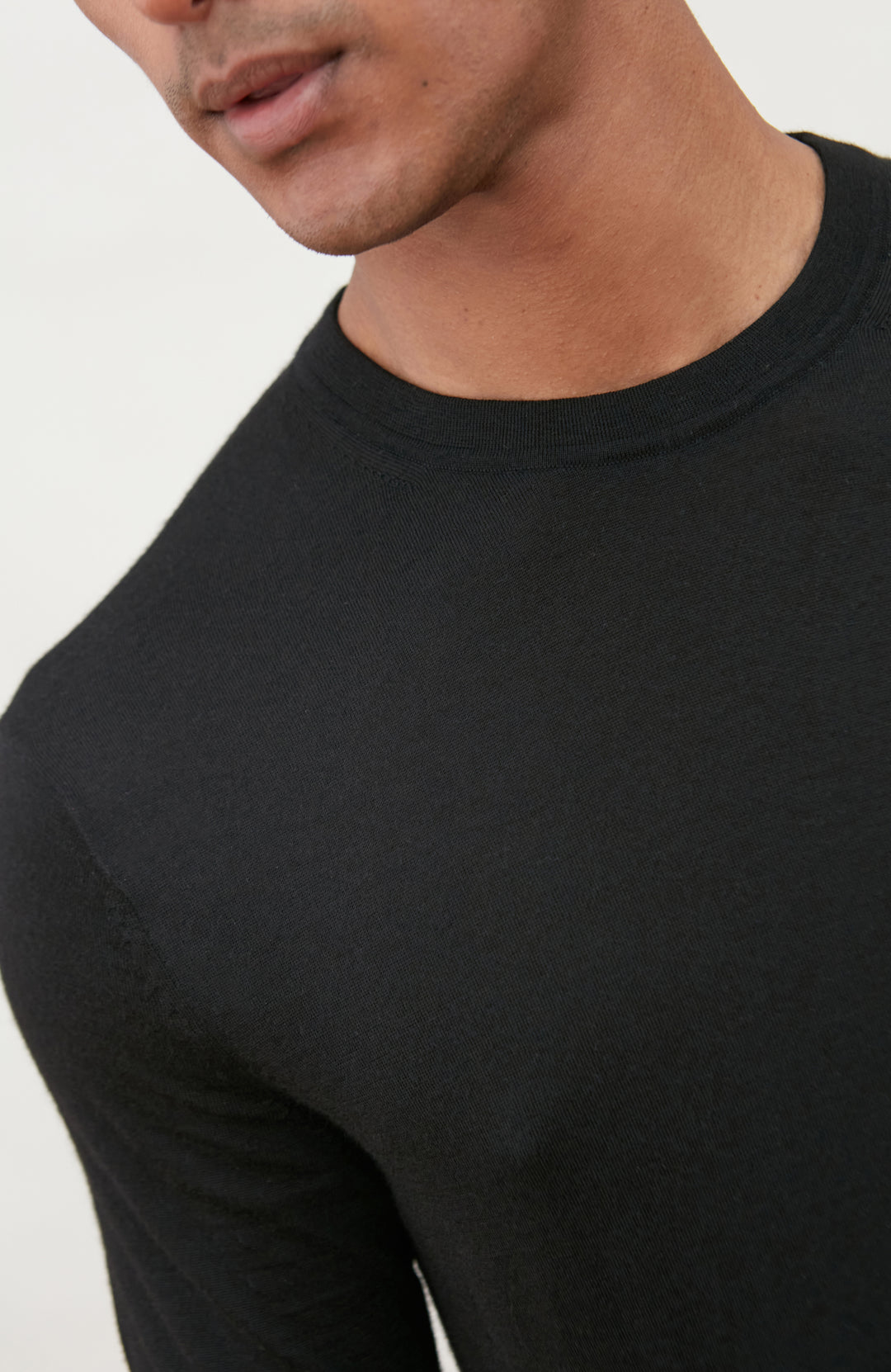 A man wearing a black short-sleeve crewneck knit sweater by Paul James Knitwear and blue jeans, standing against a light off-white background.