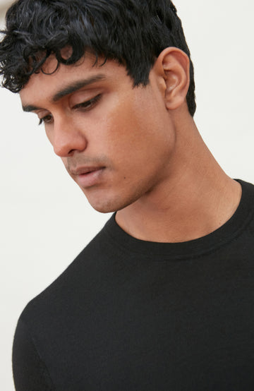 A man wearing a black short-sleeve crewneck knit sweater by Paul James Knitwear and blue jeans, standing against a light off-white background.
