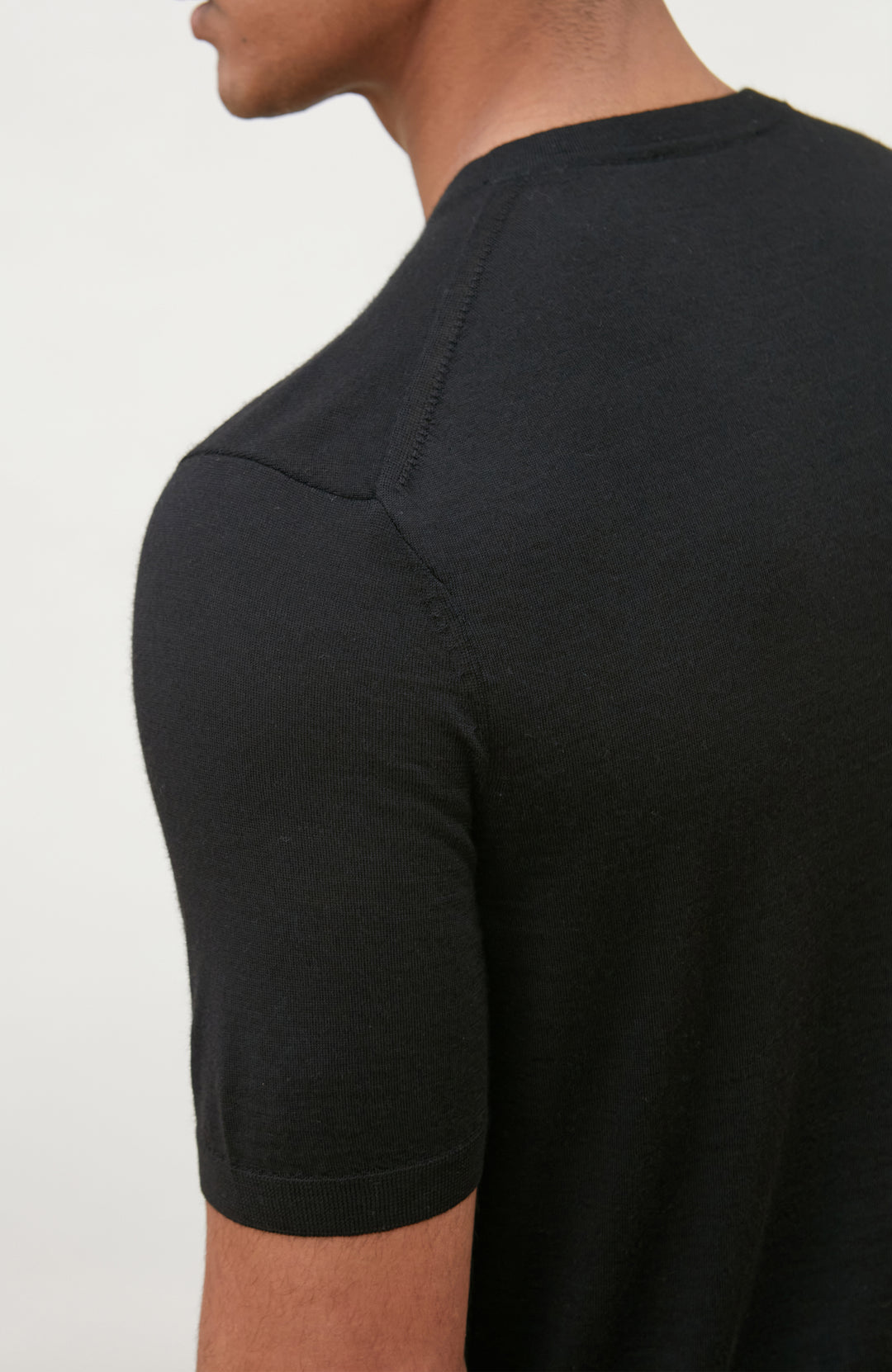 A man wearing a black short-sleeve crewneck knit sweater by Paul James Knitwear and blue jeans, standing against a light off-white background.