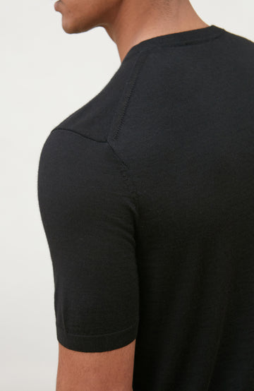 A man wearing a black short-sleeve crewneck knit sweater by Paul James Knitwear and blue jeans, standing against a light off-white background.
