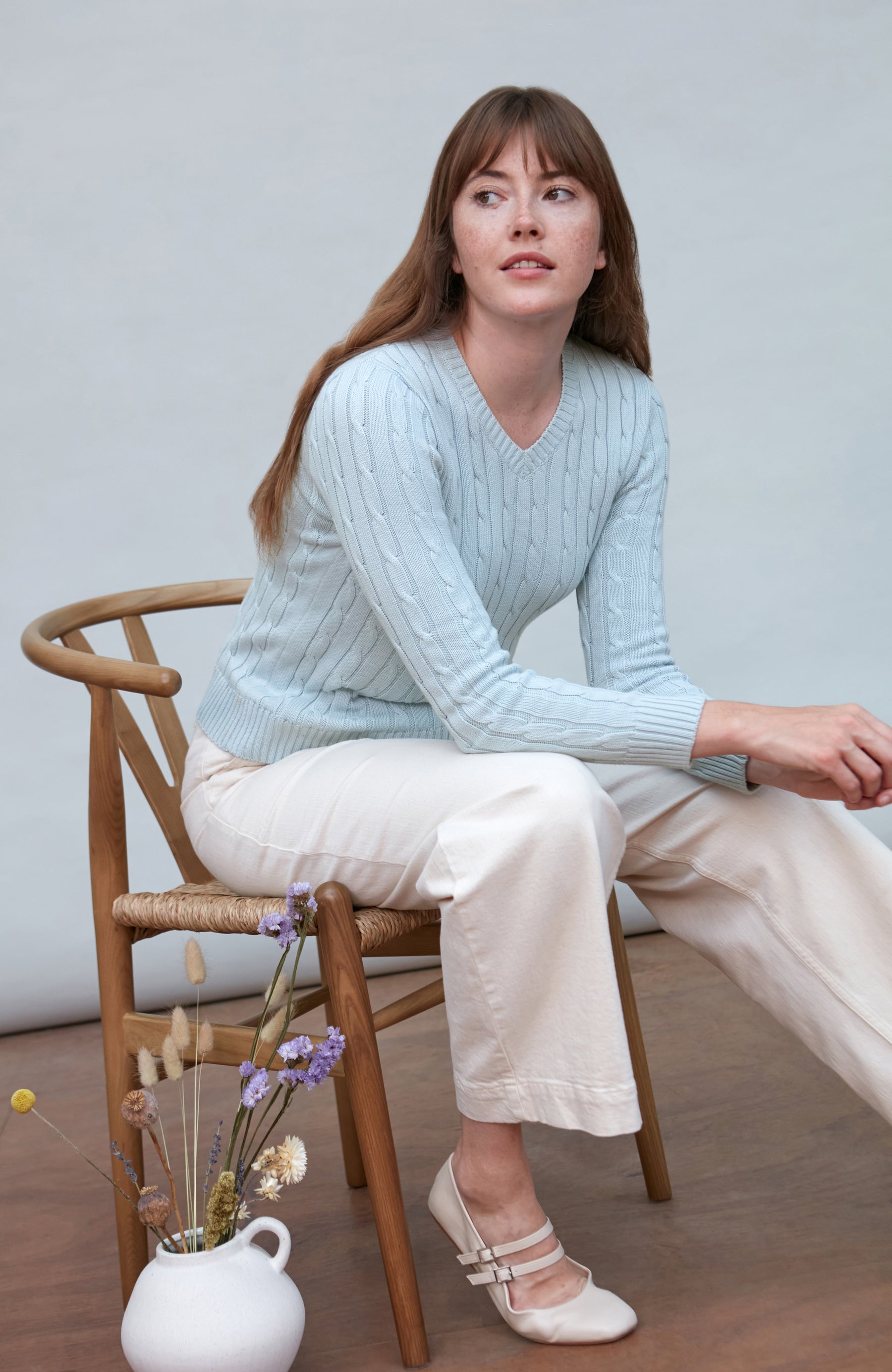 Woman sitting on a wooden chair wearing a light blue cotton cable sweater and white pants.