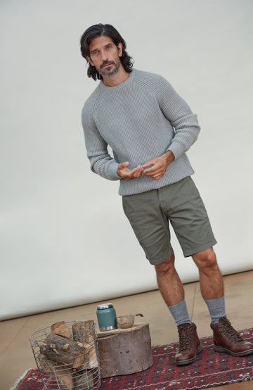 A man wearing a light gray ribbed knit Men's Ash Grey Fisherman Rib Cotton Sweater by Paul James Knitwear, olive green shorts, gray socks, and brown hiking boots, standing in a studio setting with a rug, wood slice, and wire basket on the floor.