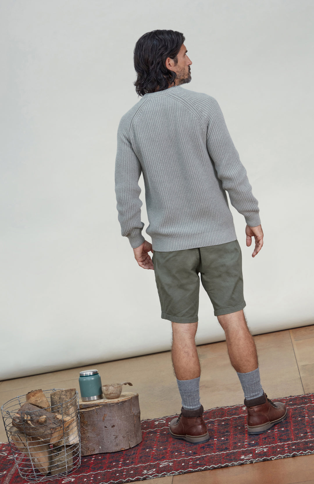 A man is shown from behind wearing a light gray ribbed knit sweater, olive green shorts, gray socks, and brown boots, standing on a patterned rug in front of a plain off-white wall. Men's Ash Grey Fisherman Rib Cotton Sweater by Paul James Knitwear.