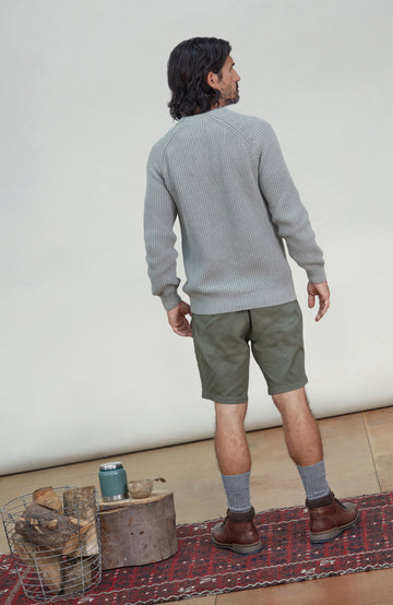 A man is shown from behind wearing a light gray ribbed knit sweater, olive green shorts, gray socks, and brown boots, standing on a patterned rug in front of a plain off-white wall. Men's Ash Grey Fisherman Rib Cotton Sweater by Paul James Knitwear.