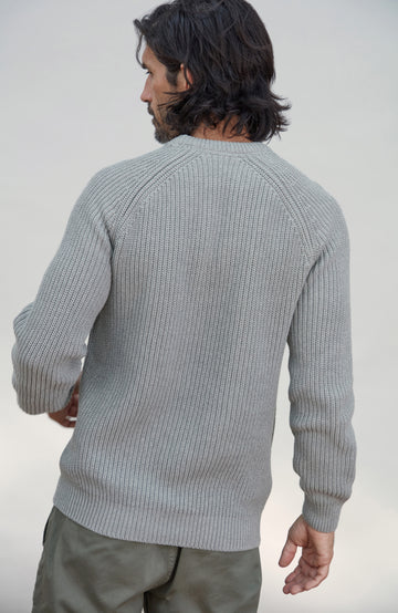 A man wearing a light gray ribbed knit sweater, shown from the back against a plain light gray studio background. Men's Ash Grey Fisherman Rib Cotton Sweater by Paul James Knitwear.