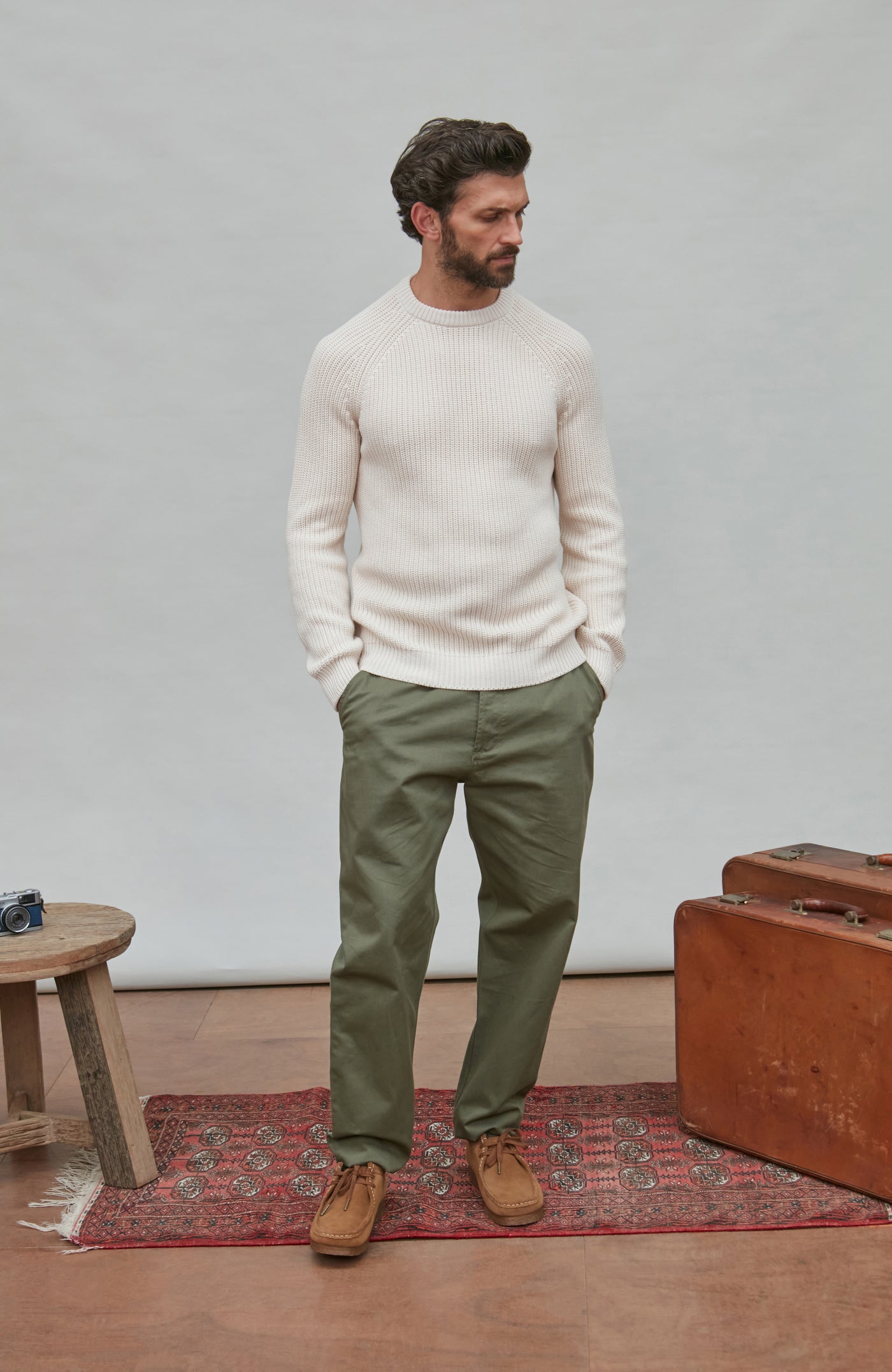 Man wearing a white fisherman rib sweater and green pants standing in a room with a suitcase and small table.