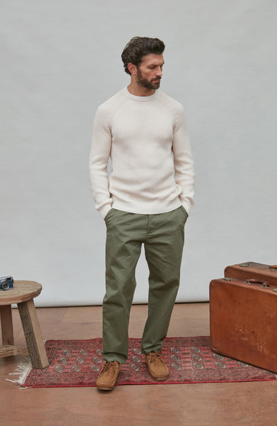 Man wearing a white fisherman rib sweater and green pants standing in a room with a suitcase and small table.