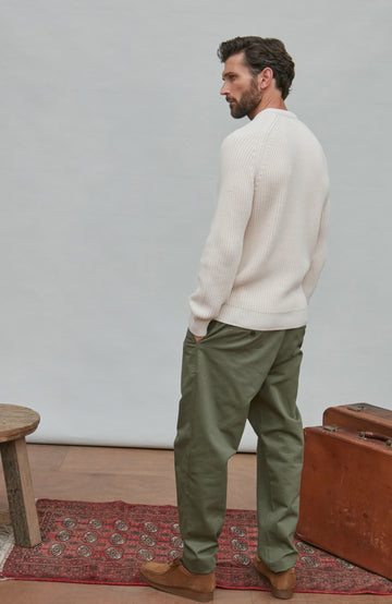 Man in a cream fisherman rib sweater and green pants standing next to a suitcase on a wooden floor.