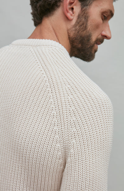 Man wearing a ecru fisherman rib knitted sweater against a plain background