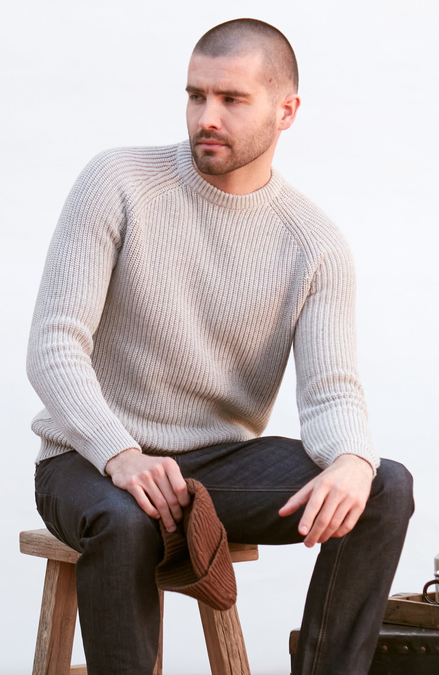 Man wearing a GREY sweater and dark pants sitting on a wooden stool against a white background
