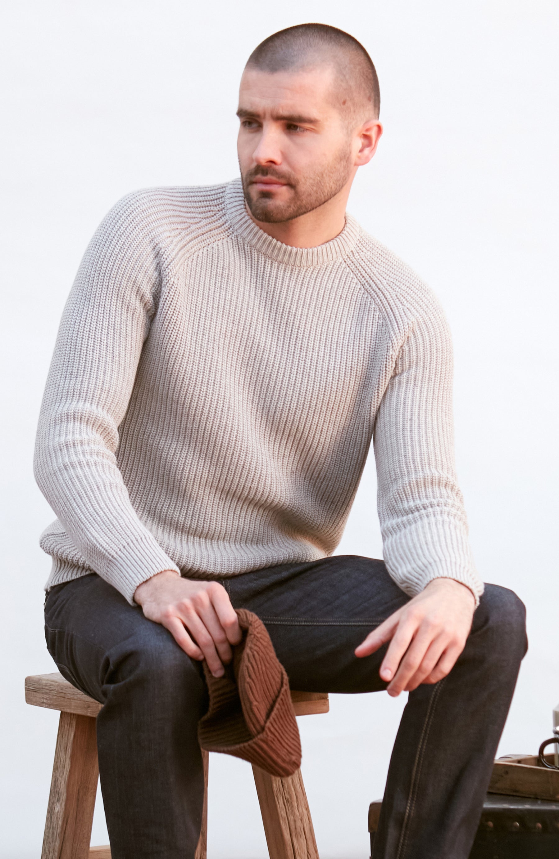 Man wearing a GREY sweater and dark pants sitting on a wooden stool against a white background