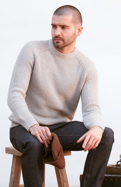 Man wearing a GREY sweater and dark pants sitting on a wooden stool against a white background