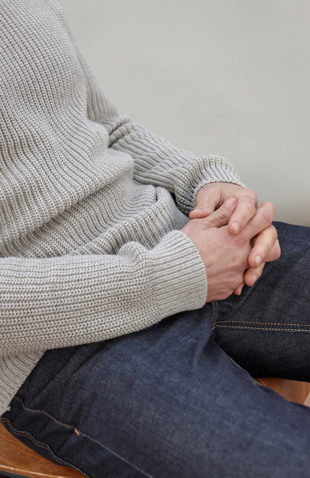 Man sitting on a chair wearing a light gray cotton fisherman sweater by Paul James Knitwear and dark jeans against a plain background