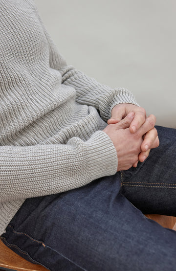 Man sitting on a chair wearing a light gray cotton fisherman sweater by Paul James Knitwear and dark jeans against a plain background