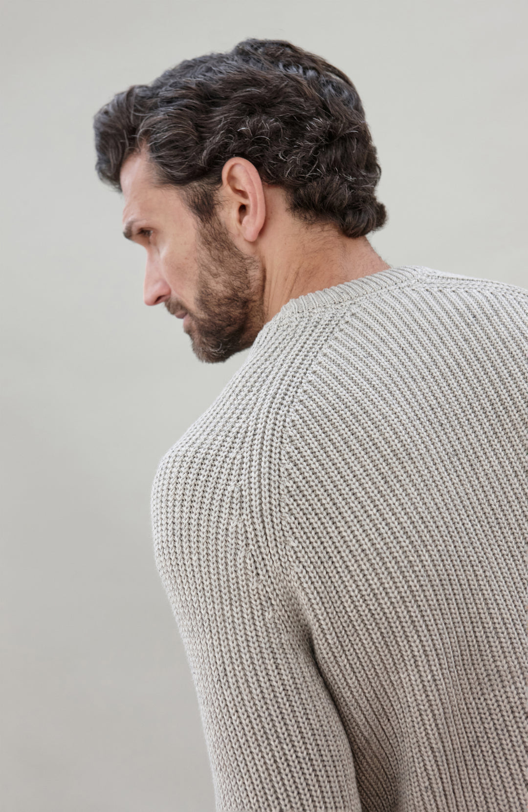 Man sitting on a chair wearing a light gray cotton fisherman sweater by Paul James Knitwear and dark jeans against a plain background