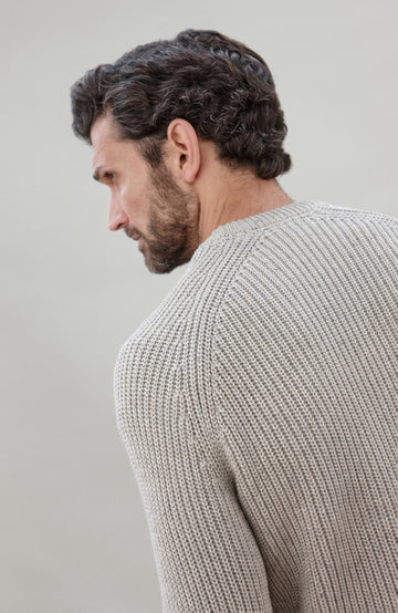 Man sitting on a chair wearing a light gray cotton fisherman sweater by Paul James Knitwear and dark jeans against a plain background