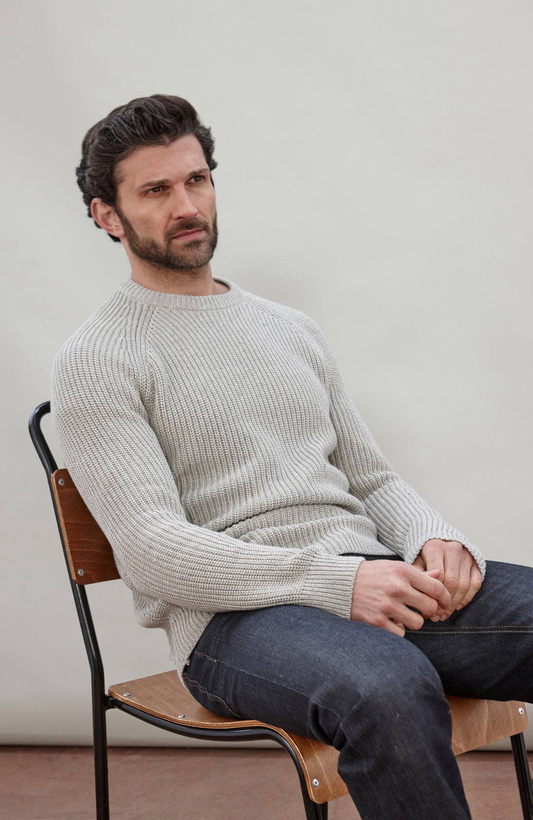 Man sitting on a chair wearing a light gray cotton fisherman sweater by Paul James Knitwear and dark jeans against a plain background