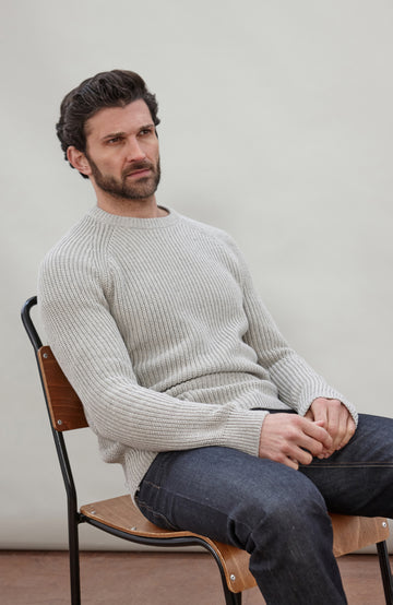 Man sitting on a chair wearing a light gray cotton fisherman sweater by Paul James Knitwear and dark jeans against a plain background